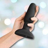 A hand holds the B-Vibe Rimming Plug 2 Vibrating Butt Plug - Black by B-Vibe against a soft, blurred background with pastel lights.